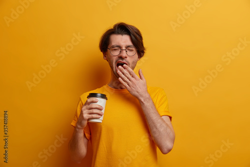 Quadro su tela Exhausted tired hipster guy yawns and wants to sleep, drinks refreshing beverage from disposable cup, needs rest, wears glasses and yellow t shirt, awakes very early