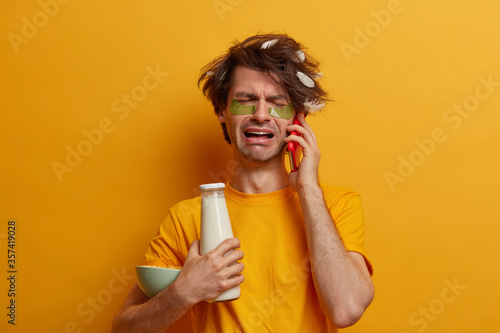 Morning time, daily routine concept. Desperate miserable man with feather on hair, cries while talks on cellular, holds cereals with milk for breakfast, undergoes beauty treatments, reduces wrinkles © wayhome.studio 