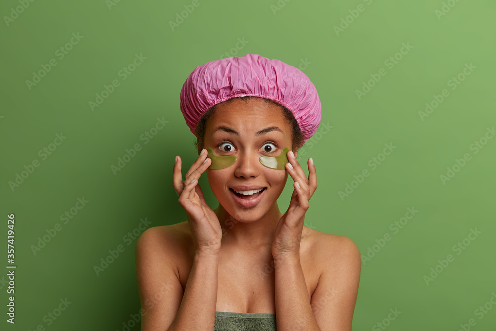 Portrait of surprised dark skinned female touches soft skin, applies eye patches for reducing puffiness and fine lines, wears bath hat, soft towel around body, isolated over green background. Portrait of surprised dark skinned female touches soft skin, applies eye patches for reducing puffiness and fine lines, wears bath hat, soft towel around body, isolated over green background.