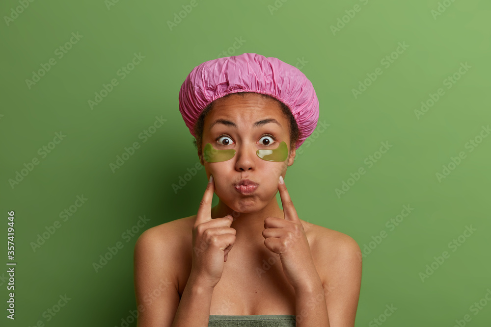 Cosmetology, skin care and facial treatment concept. Half naked dark skinned woman blows cheeks, applies collagen patches under eyes for reducing puffiness, enjoys spa procedures, wears bath hat Cosmetology, skin care and facial treatment concept. Half naked dark skinned woman blows cheeks, applies collagen patches under eyes for reducing puffiness, enjoys spa procedures, wears bath hat