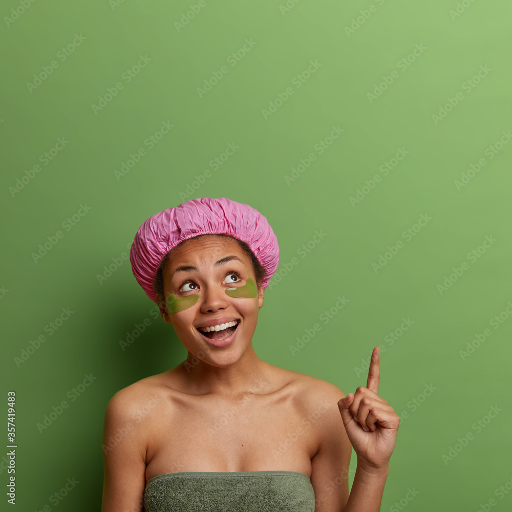 Happy young female model points above with fore finger, recommends cosmetology product for healthy skin, wears bath hat and green patches under eyes, feels positive, poses indoor, empty space up Happy young female model points above with fore finger, recommends cosmetology product for healthy skin, wears bath hat and green patches under eyes, feels positive, poses indoor, empty space up