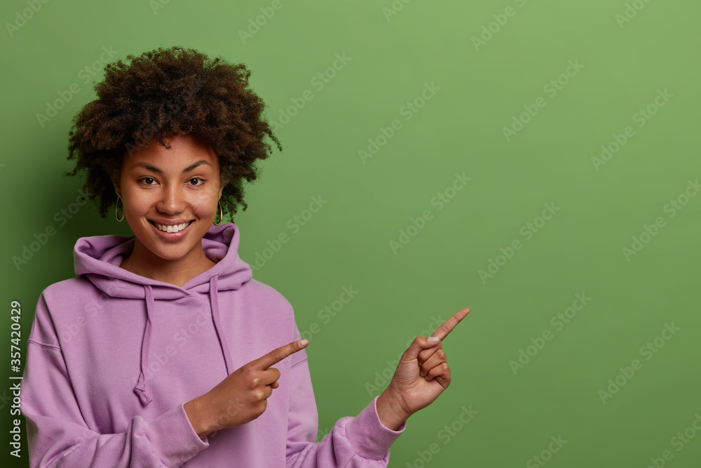Positive curly haired millennial girl in casual purple hoodie points aside on empty copy space for your advertisement, being in good mood, isolated over green wall, shows way to best online courses Positive curly haired millennial girl in casual purple hoodie points aside on empty copy space for your advertisement, being in good mood, isolated over green wall, shows way to best online courses