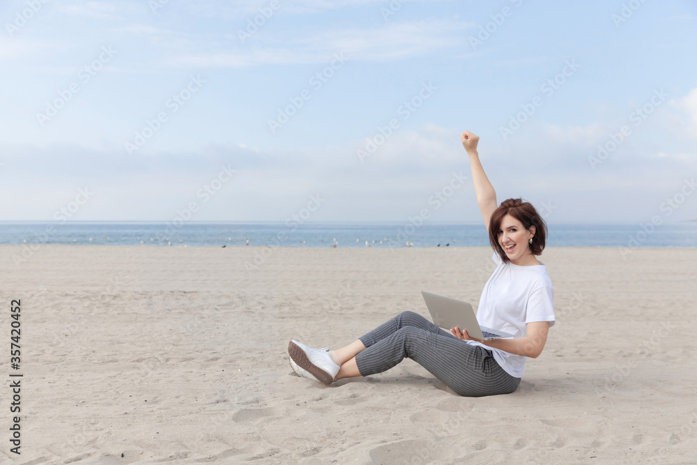 Happy freelance woman raised her hands up while sitting on the empty beach / sand with laptop. 