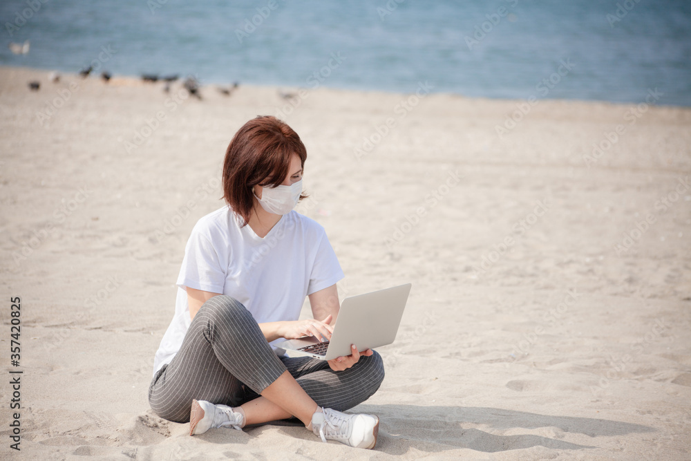 Young woman in medical mask sitting with laptop on empty coast, protective equipment during coronavirus. Freelance concept, working on the beach. Distance education on the beach with medical mask.