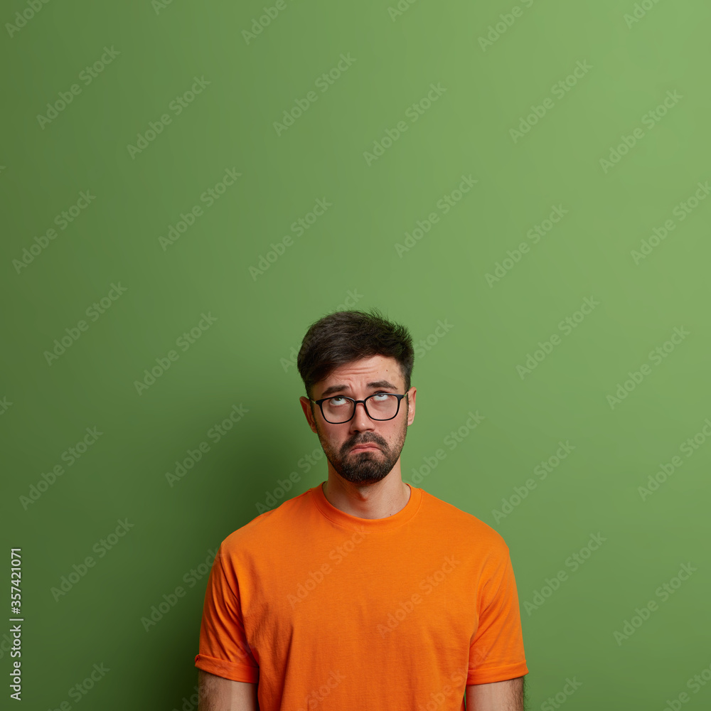 Gloomy dissatisfied man looks upwards, smirks face and stays serious, displeased with something unfair, wears glasses and casual clothing, isolated on green background, blank space for advert Gloomy dissatisfied man looks upwards, smirks face and stays serious, displeased with something unfair, wears glasses and casual clothing, isolated on green background, blank space for advert