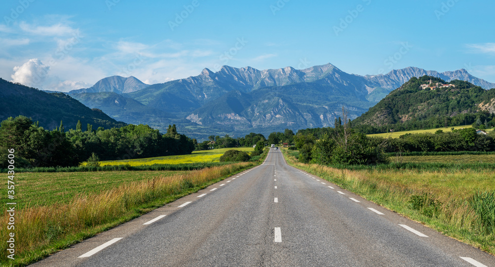 A long straight road leading towards a mountains in France. Amazing bright colorful spring and summer landscape. Yellow fields of flowering rape and blue sky with clouds. 