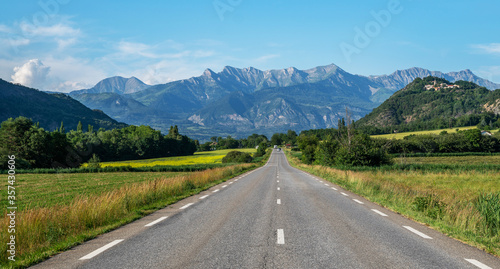 A long straight road leading towards a mountains in France. Amazing bright colorful spring and summer landscape. Yellow fields of flowering rape and blue sky with clouds. 