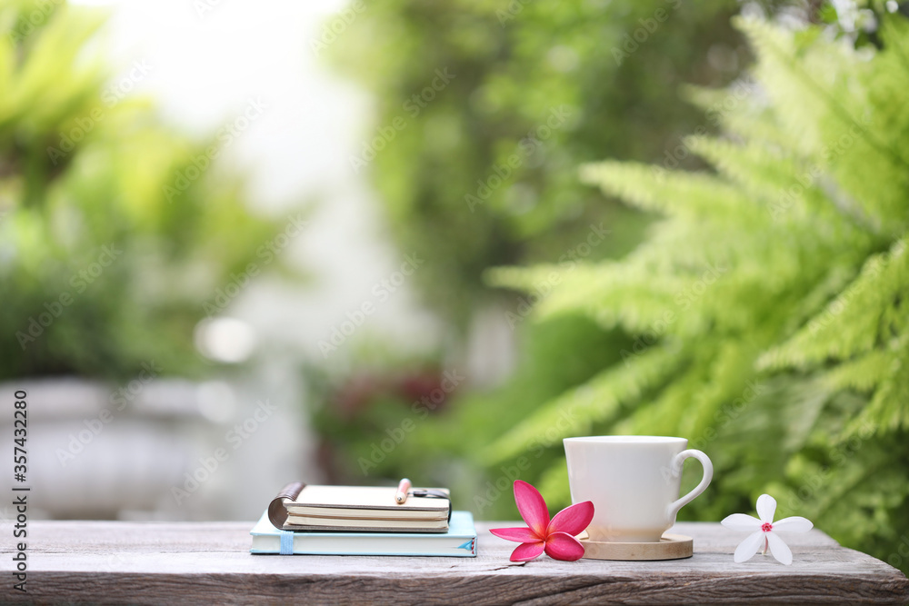 White coffee cup and notebooks with flowers on brown wooden table at outdoor lush scenery White coffee cup and notebooks with flowers on brown wooden table at outdoor lush scenery