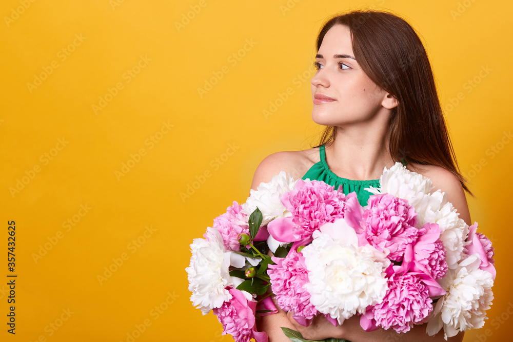 Profile of brunette young lady wearing green clothing, embracing huge bouquet of peonies, female looking aside, standing against yellow background. Copy space for advertisement or promotional text.