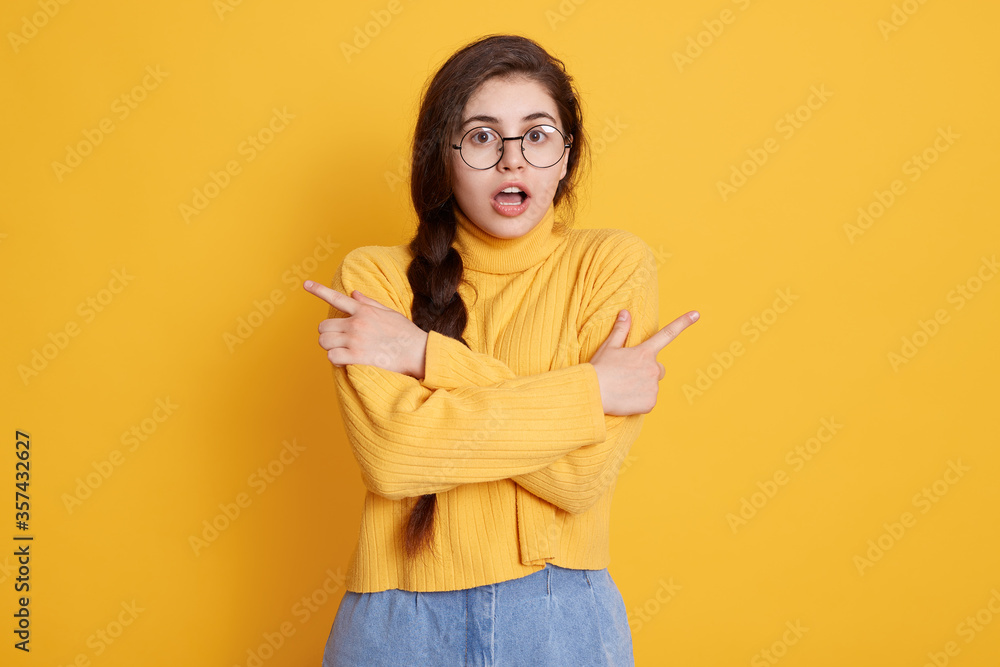 Surprised female wearing stylish clothing pointing with index fingers to sides, standing isolated over yellow background, brunette girl stands with opened mouth. Surprised female wearing stylish clothing pointing with index fingers to sides, standing isolated over yellow background, brunette girl stands with opened mouth.