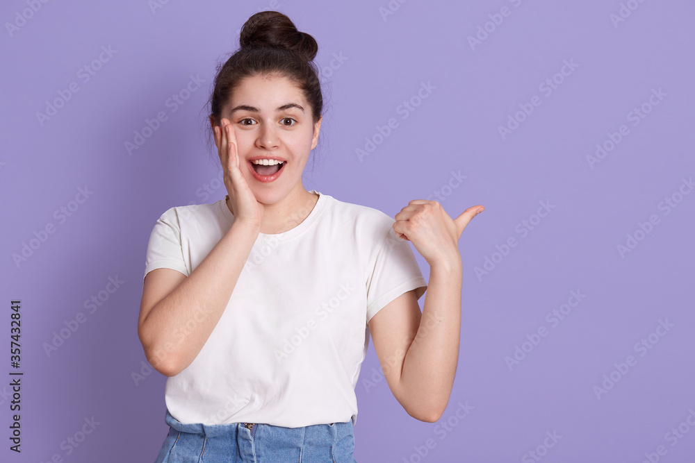 Excited smiling happy woman touching her cheek with palm and pointing aside with thumb, showing something amazement, keeps mouth opened, wearing white t shirt. Excited smiling happy woman touching her cheek with palm and pointing aside with thumb, showing something amazement, keeps mouth opened, wearing white t shirt.