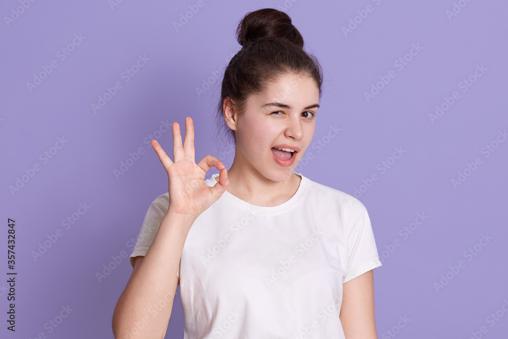 Young adorable female with dark hair showing ok sign with fingers, winks with her eye, woman has hair bun, dresses white casual t shirt, posing against lilac background. Young adorable female with dark hair showing ok sign with fingers, winks with her eye, woman has hair bun, dresses white casual t shirt, posing against lilac background.