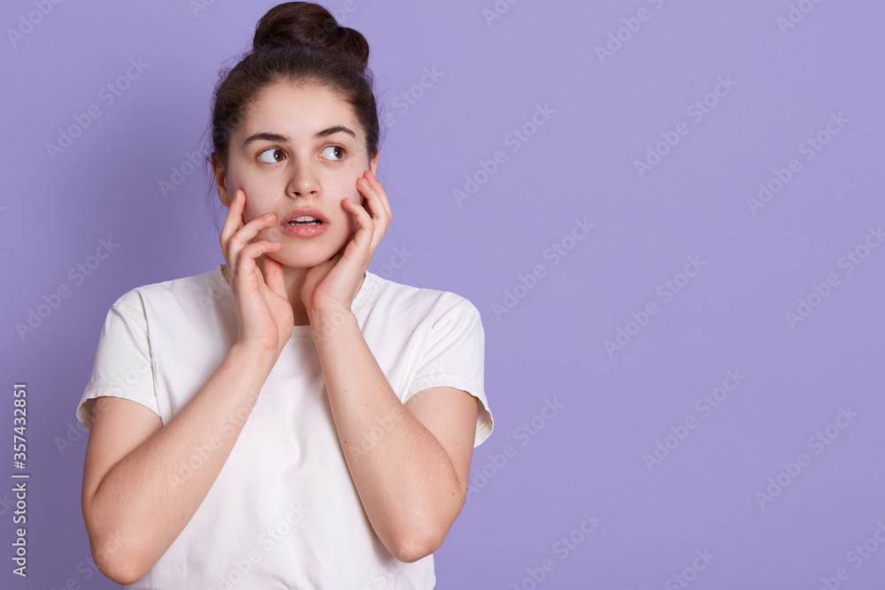 Beautiful female with hair bun looking aside with astonished facial expression, touching her cheeks with palm, wearing white t shirt. Copy space for advertisement. Beautiful female with hair bun looking aside with astonished facial expression, touching her cheeks with palm, wearing white t shirt. Copy space for advertisement.
