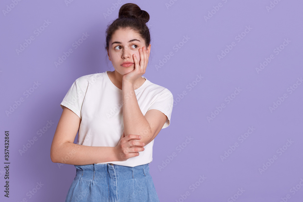 Brunette woman standing isolated over lilac background, caucasian lady with her hand under chin looking aside with thoughtful facial expression, female with hair bun, dresses casually. Brunette woman standing isolated over lilac background, caucasian lady with her hand under chin looking aside with thoughtful facial expression, female with hair bun, dresses casually.