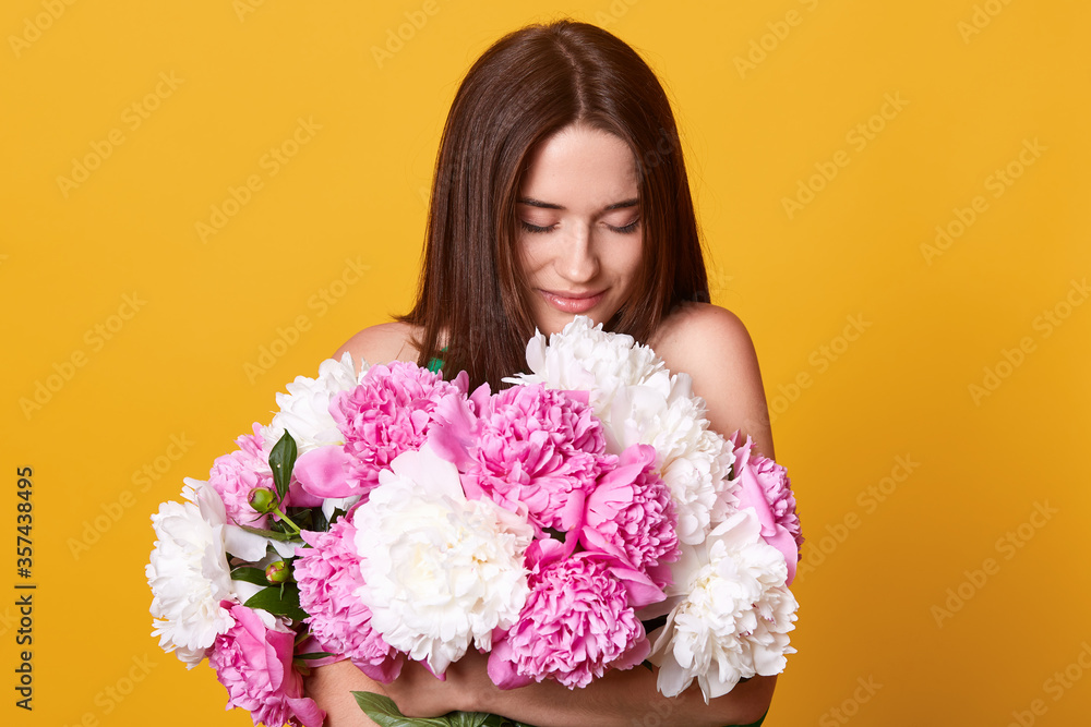 Brunette gentle lady smells peony flowers, dark haired girl enjoying beautiful smell of flower, female looks down on her bouquet, standing against yellow background.