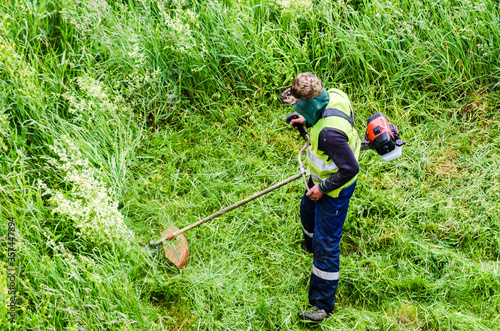 Worker mower in protective coveralls mows the tall grass with a mechanical lawn mower in a meadow