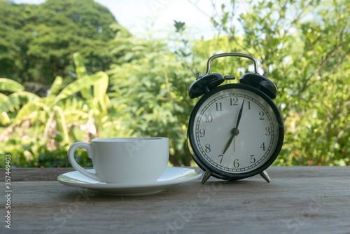 A white cup and a vintage alarm clock on wooden background on a bright day with a nature background.
