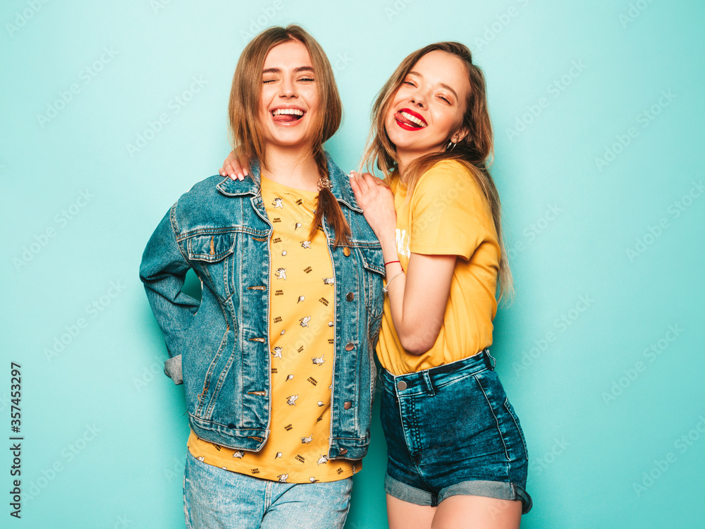Two young beautiful smiling hipster girls in trendy summer yellow T-shirts and jeans jacket. Sexy carefree women posing near blue wall. Trendy and positive models having fun Two young beautiful smiling hipster girls in trendy summer yellow T-shirts and jeans jacket. Sexy carefree women posing near blue wall. Trendy and positive models having fun
