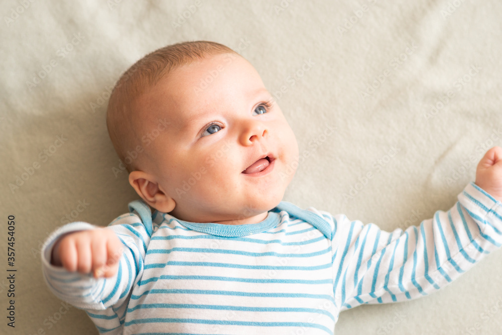 Happy, amused baby boy in blue striped bodysuit lying down on grey blanket, smiling to someone , something on right side. Three months old, blue-eyed caucasian baby with brown hair. Happy, amused baby boy in blue striped bodysuit lying down on grey blanket, smiling to someone , something on right side. Three months old, blue-eyed caucasian baby with brown hair.