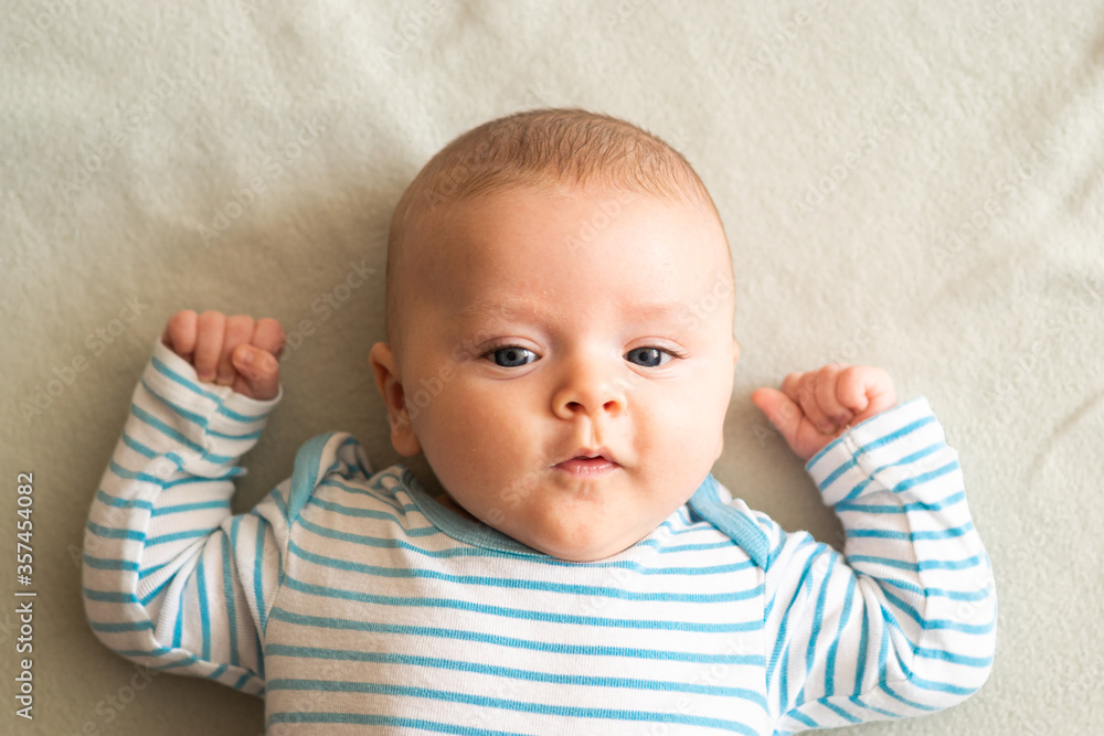 Surprised, focused caucasian baby boy looking straight to the camera, wearing blue striped clothes, lying down on grey blanket