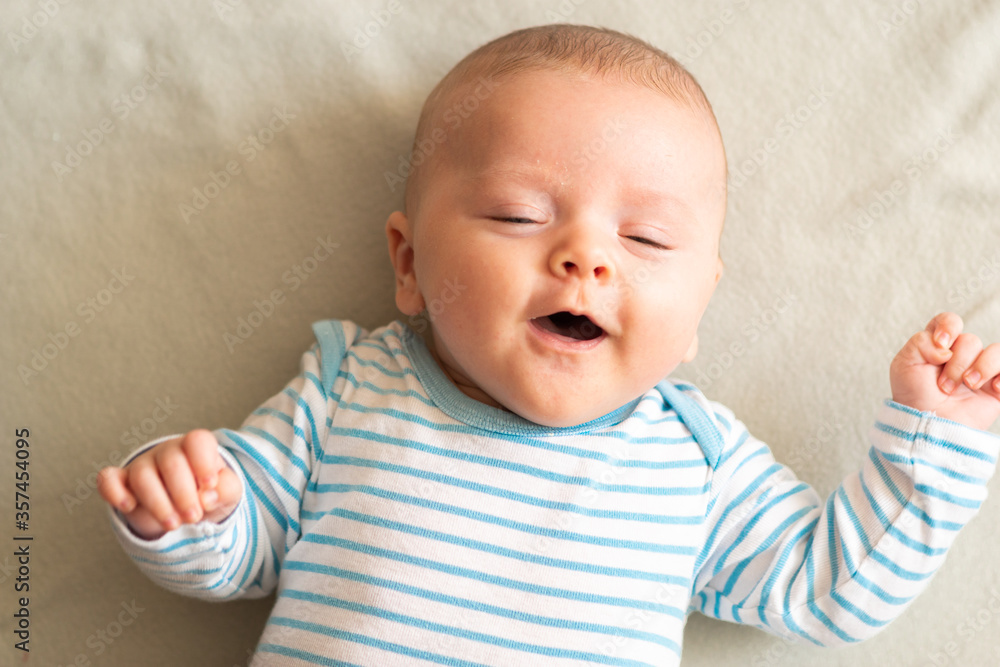 Funny little 3 months old baby boy laughing and smiling with his eyes closed, lying down on grey blanket. Happy toddler in blue striped bodysuit.