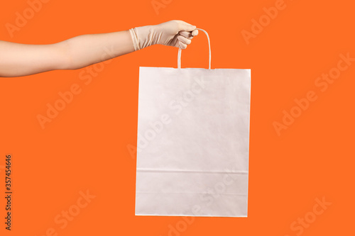 Profile side view closeup of human hand in white surgical gloves holding and showing white shopping bag. indoor, studio shot, isolated on orange background.