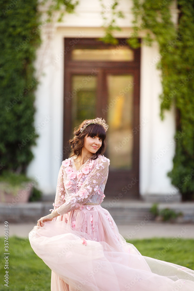 Fototapeta premium Beautiful woman in flying airy pink dress on background of door overgrown with wild grapes. Pretty young caucasian girl plays with her dress. Girl dance in flowing dress