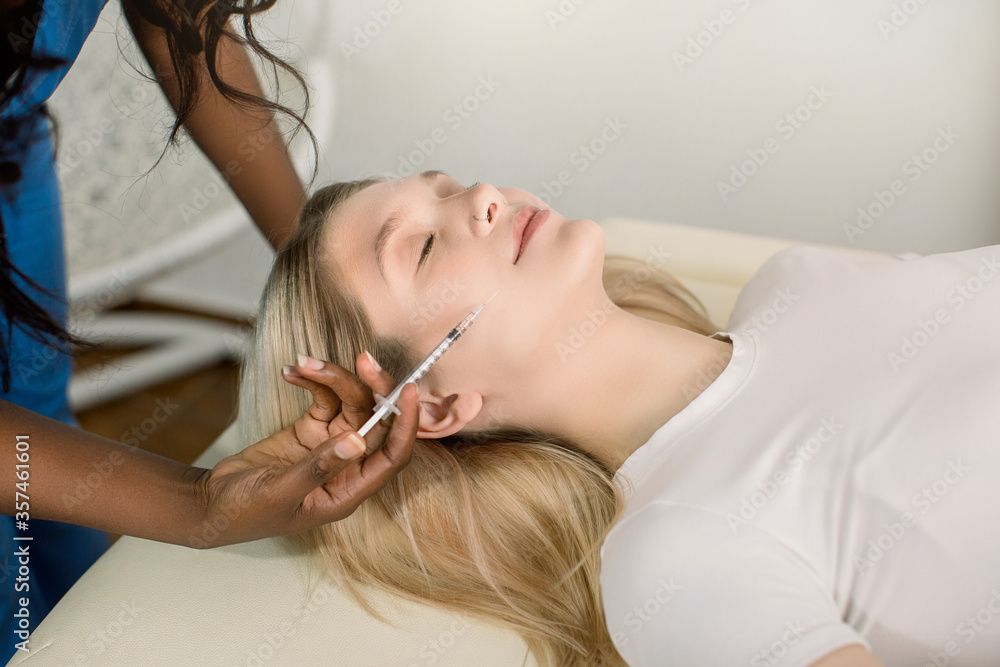 Pretty young Caucasian lady lying on the couch and receiving beauty injections. African dark skinned beautician makes anti-aging injection on woman face. Rejuvenation and beauty treatments. Pretty young Caucasian lady lying on the couch and receiving beauty injections. African dark skinned beautician makes anti-aging injection on woman face. Rejuvenation and beauty treatments.