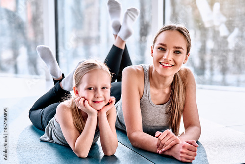 Beautiful young woman and charming little girl are looking at camera and smiling while lying on yoga mat in fitness hall photo