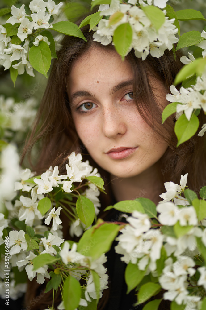 A large portrait of a brunette girl standing next to a tree in white Apple blossoms and looking directly at the camera A large portrait of a brunette girl standing next to a tree in white Apple blossoms and looking directly at the camera