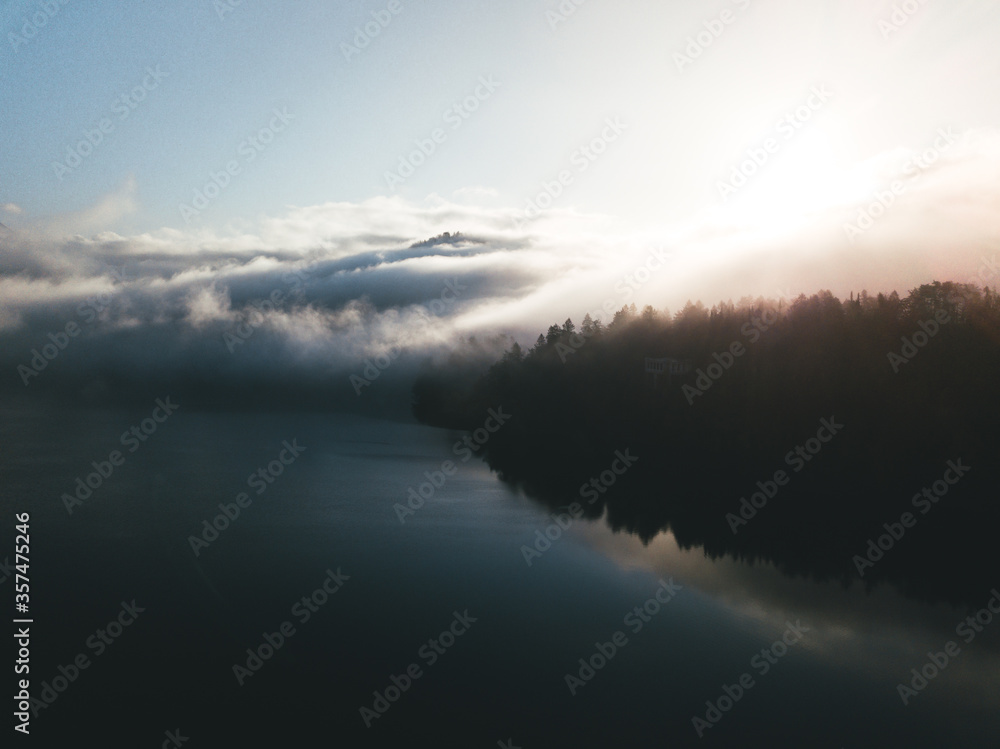 Fototapeta premium Moody scenic view of lake against sky. Amazing sunrise above lake and forest with low fog and clouds surrounding the mountains. Serene moment and fantastic scenery at lake Bled, Slovenia