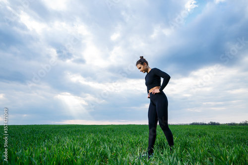 Young attractive brunette woman standing in nature, wearing black leggings and a black top. Summer day, green grass. Athletic body and healthy lifestyle. © Дмитрий Ткачук