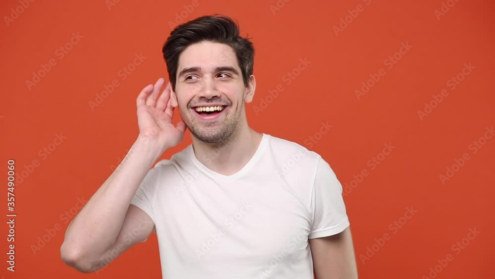 Young fun guy 20s 30s years old in white t-shirt isolated ovn orange wall background studio. People sincere emotions lifestyle concept. Looking aside try to hear you with hand at ear overhears secrets Young fun guy 20s 30s years old in white t-shirt isolated ovn orange wall background studio. People sincere emotions lifestyle concept. Looking aside try to hear you with hand at ear overhears secrets