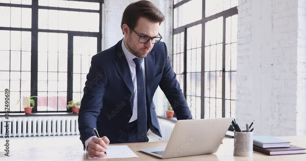 Smart skilled young male executive wearing suit, working on computer, managing tasks standing at workplace. Busy 30s businessman developing company growth strategy, analyzing financial market online. Smart skilled young male executive wearing suit, working on computer, managing tasks standing at workplace. Busy 30s businessman developing company growth strategy, analyzing financial market online.