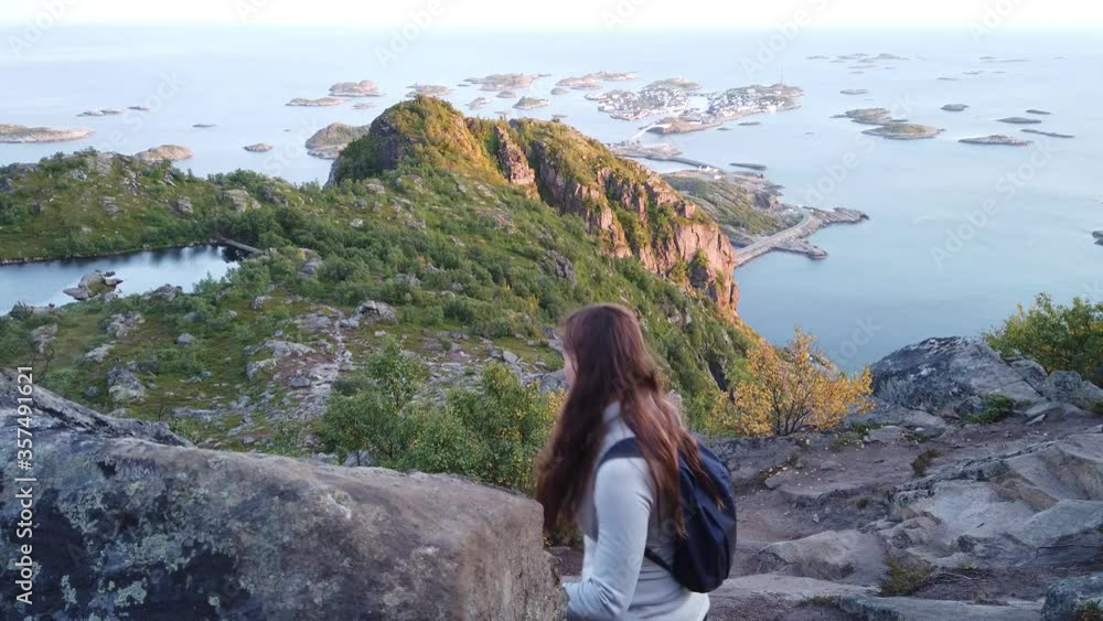 A young brunette woman is being careful and balancing her steps down the mountain on the dirt trail between big rocks and stones. Beautiful lake and islands in the background. Henningsvaer, Lofoten. A young brunette woman is being careful and balancing her steps down the mountain on the dirt trail between big rocks and stones. Beautiful lake and islands in the background. Henningsvaer, Lofoten.