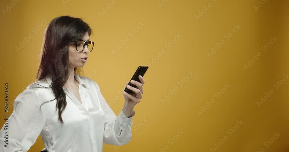 Asian girl in glasses surfing internet on smatphone then pointing in air, gesturing thumb up, looking at camera and smiling, isolated on yellow background close up 4k Asian girl in glasses surfing internet on smatphone then pointing in air, gesturing thumb up, looking at camera and smiling, isolated on yellow background close up 4k