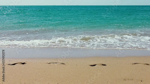 A man walks along a sandy beach leaving footprints in the sand , which are slowly washed away by the turquoise sea waves.