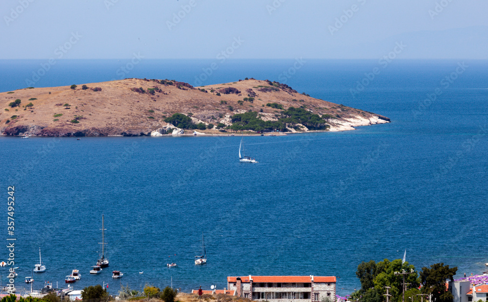 Aerial shot of a small sailboat sailing before the small island in Foca, Izmir, Turkey. People are enjoying the sea at the beaches swimming and barbecuing.
