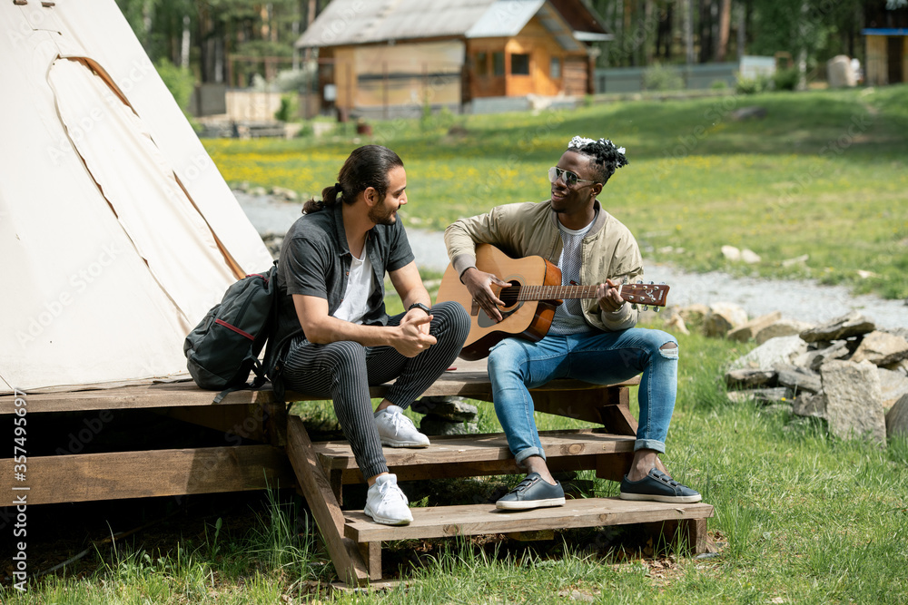 Black guy sitting on step and playing guitar for friend while they spending time at countryside festival