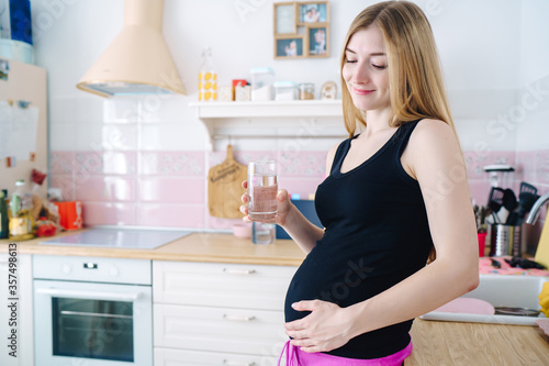 young beautiful woman drinks a glass of water in the kitchen and smiles and looks at the pregnant belly, close-up, concept of a healthy lifestyle, water balance