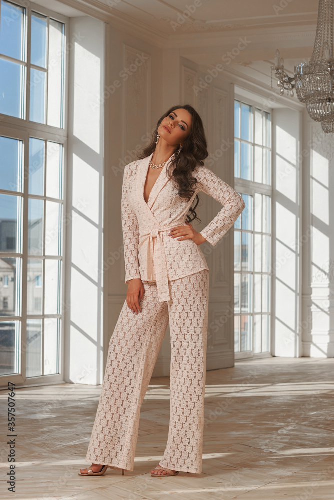 Fototapeta premium Young beautiful woman with long brown wavy hair in beige lace suit standing and posing in luxury interior with french windows on a sunny day. Interior fashion portrait with beautiful light and shadows