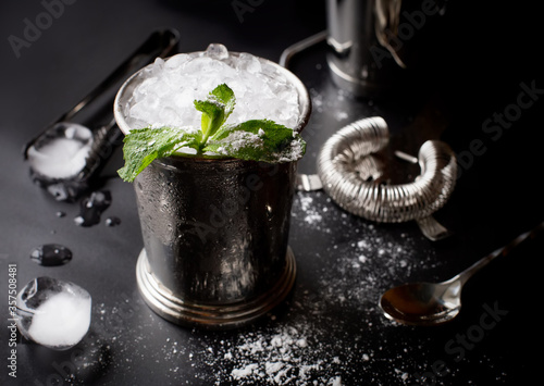 Fotografie Mint julep whiskey cocktail in misted frozen metal glass and bar accessories shaker jigger spoon ice sugar on black background