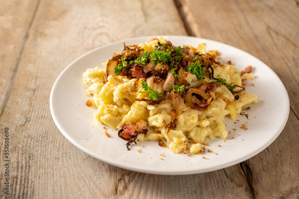 Homemade spaetzle, German egg noodles with cheese, roasted onions, bacon and breadcrumbs, served with parsley garnish on a white plate on a rustic wooden table, selected focus