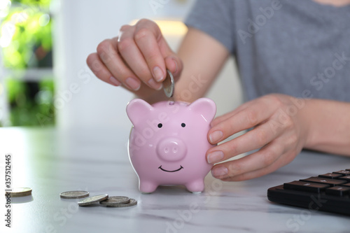 Woman putting money into piggy bank at marble table indoors, closeup