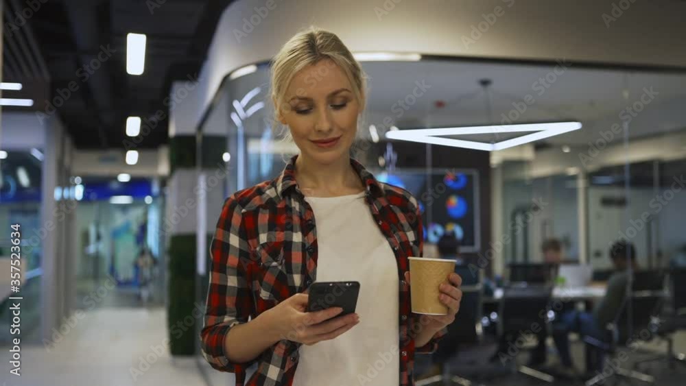 Medium waist up shot of young blonde woman in checkered shirt walking down office corridor in slow motion and text messaging on cell phone, disposable coffee cup in her hand Medium waist up shot of young blonde woman in checkered shirt walking down office corridor in slow motion and text messaging on cell phone, disposable coffee cup in her hand