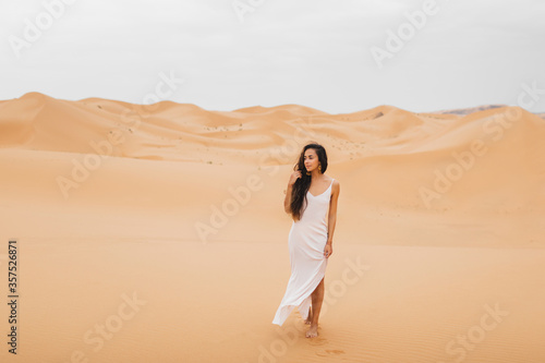 Beautiful woman in gentle silk dress walking in sand dunes of Morocco Sahara desert. Brunette with long hair, eastern appearance.