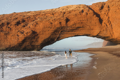 Couple in love walking under huge arch on Legzira ocean beach at sunset. Famous and popular landmark in Morocco. photo