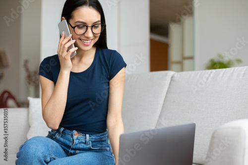 Cheerful Girl Chatting On Cellphone Sitting At Laptop At Home