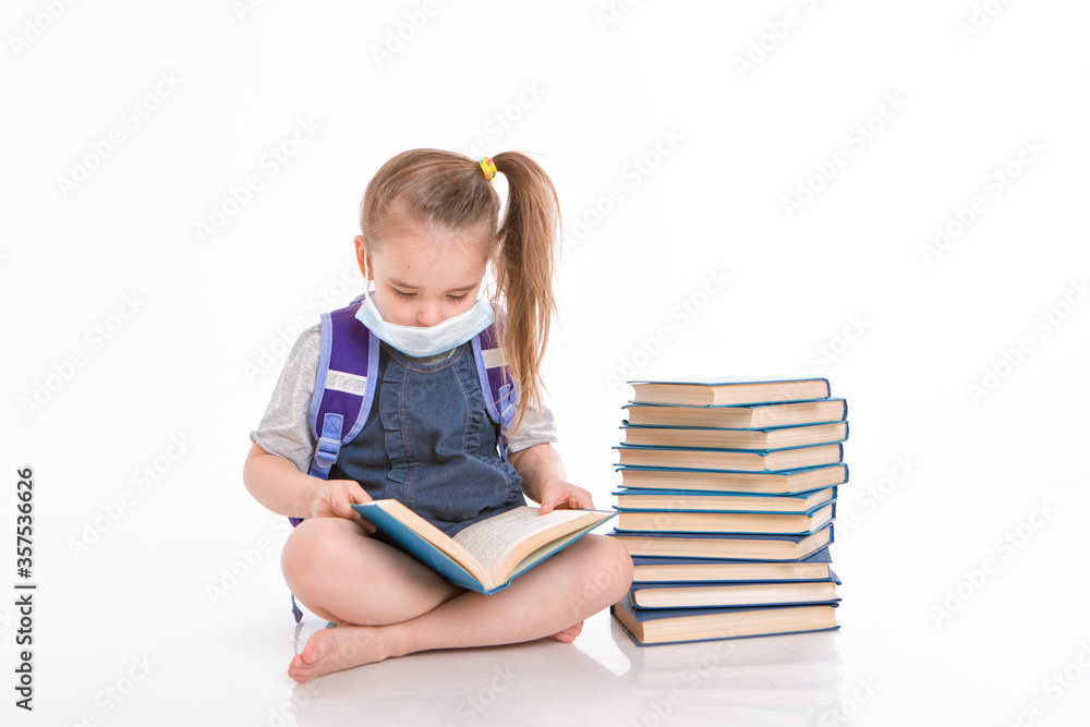 a first-grader learns to read. a little girl on home distance learning. A child in a medical mask reads a book. the student is doing homework. isolated on a white background a first-grader learns to read. a little girl on home distance learning. A child in a medical mask reads a book. the student is doing homework. isolated on a white background