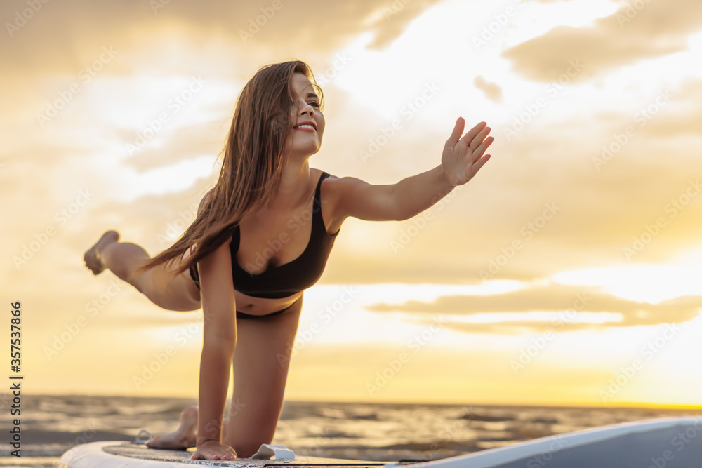 Young sexy woman surfer doing yoga on her sup board on the sunset. Rest at nature concept